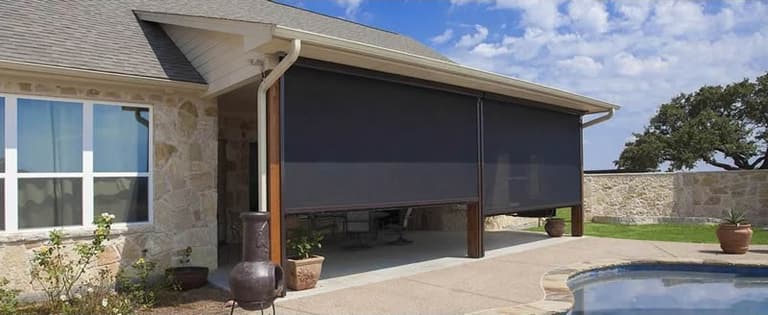 Two motorized solar screens lowered on a stone home’s covered patio next to a pool